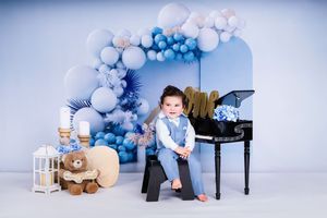 First birthday photo setup with blue and white balloons, teddy bears, and piano decorations.
