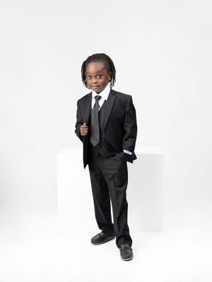 Young child wearing a black suit and tie gives thumbs up against white background.