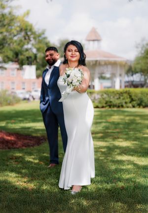Bride and groom poses in a grassy outdoor setting with trees and brick buildings in the background.