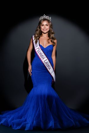 Pageant contestant in royal blue evening gown poses with crown and sash against dark background.