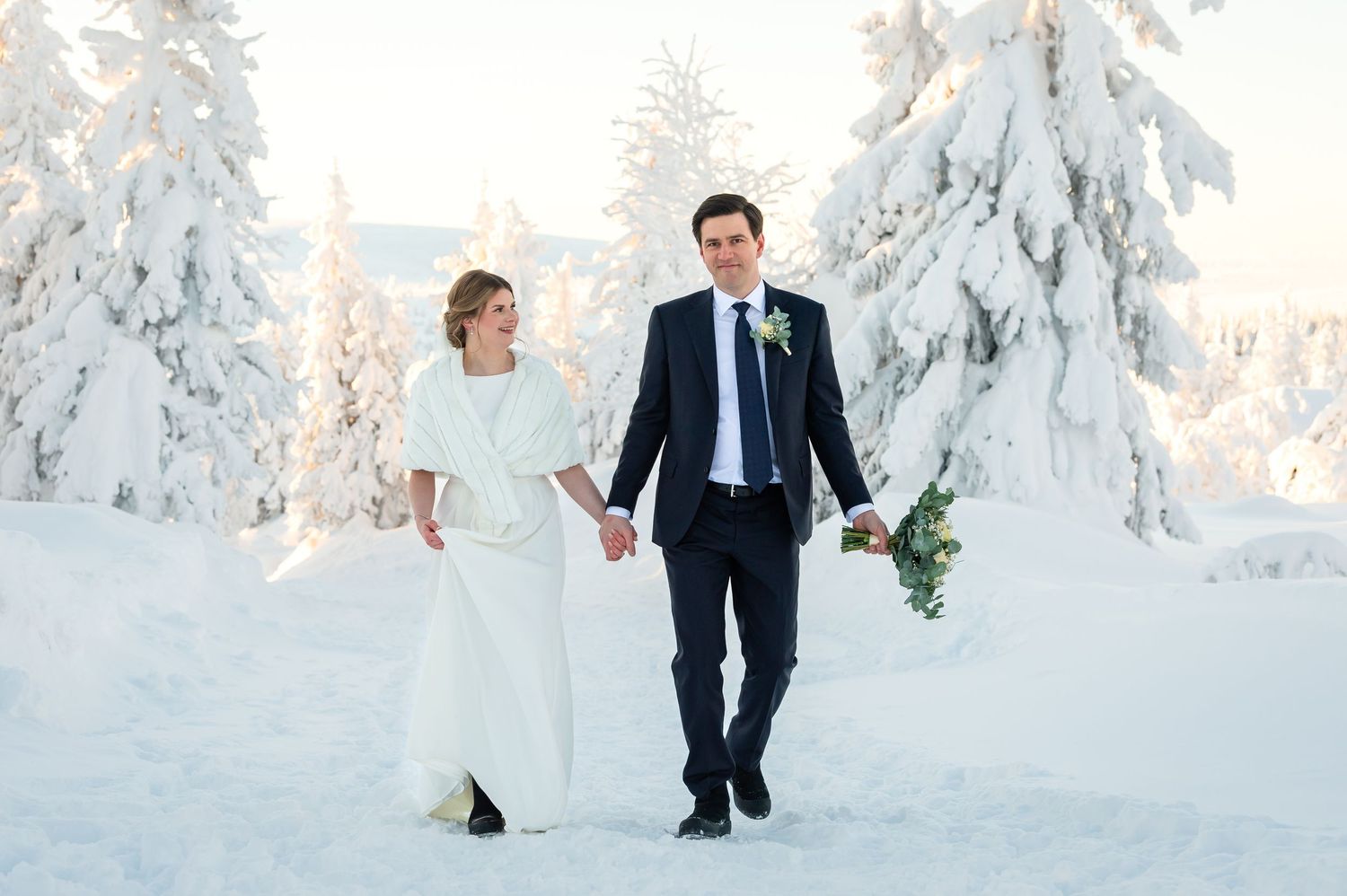 Brudpar kommer gående mellan snöklädda granar, brudgum håller brudbukett och bruden har en pälsbolero och svarta skor