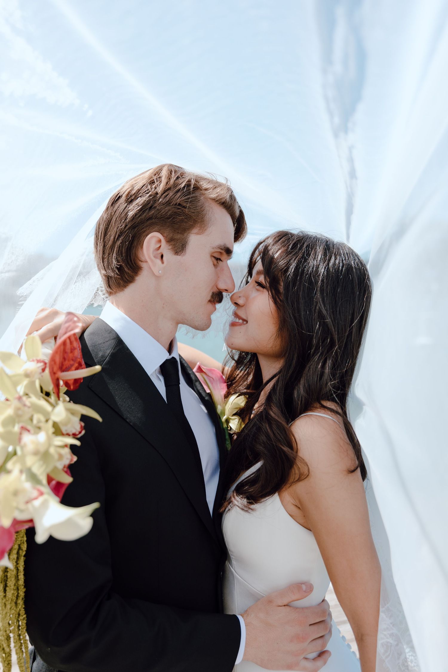 A newlywed couple shares a romantic kiss in a lush garden setting while holding a bridal bouquet.