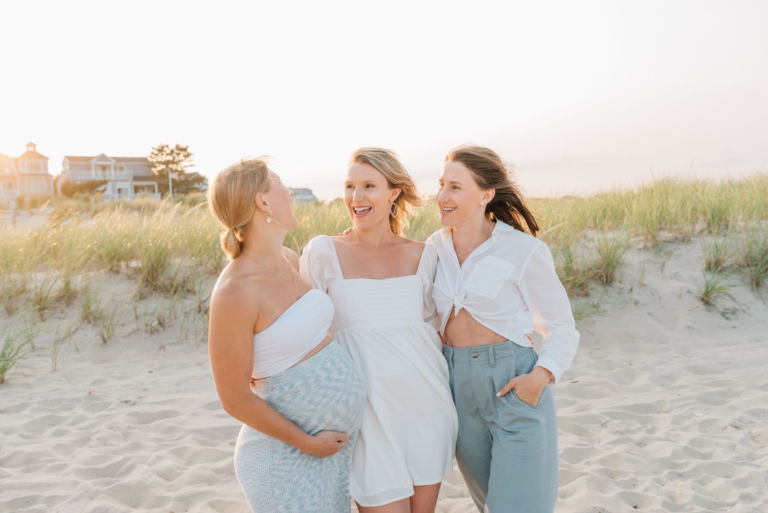 Three sisters laugh on the beach in summer outfits.