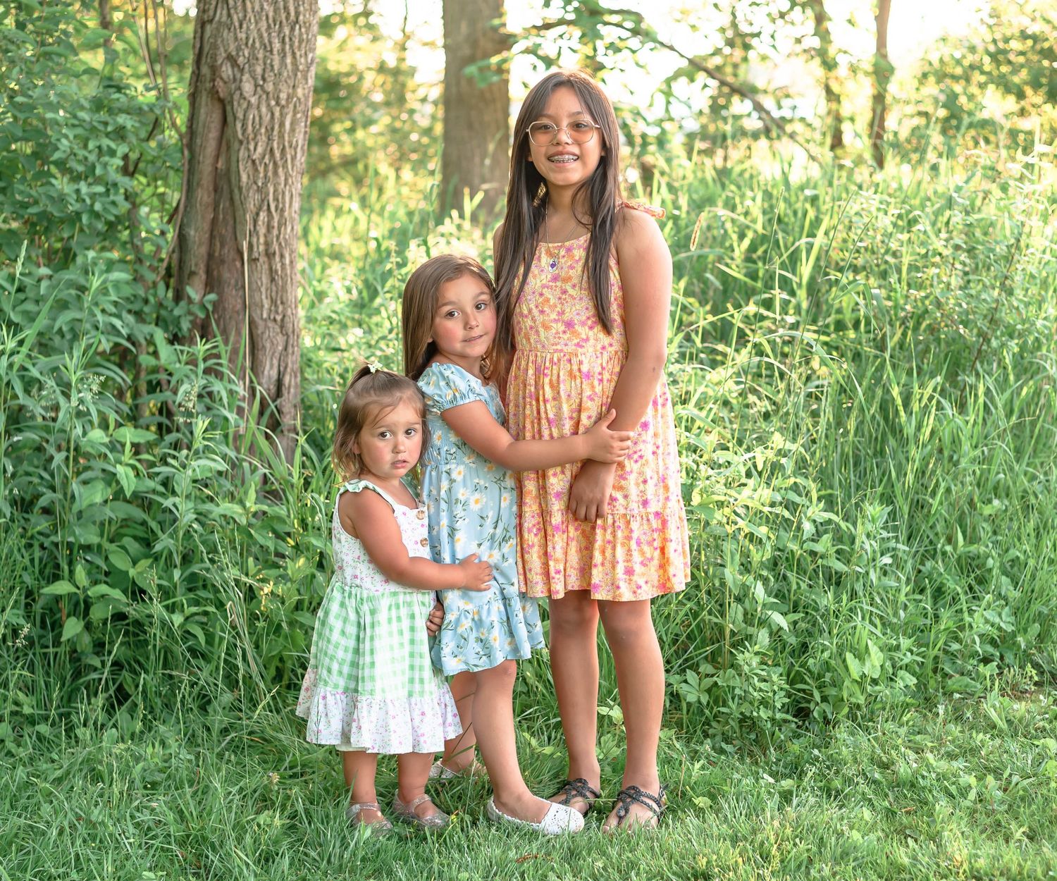 Three sisters in summer dresses stand together in front of a tree surrounded by tall grass in a sunlit forest.