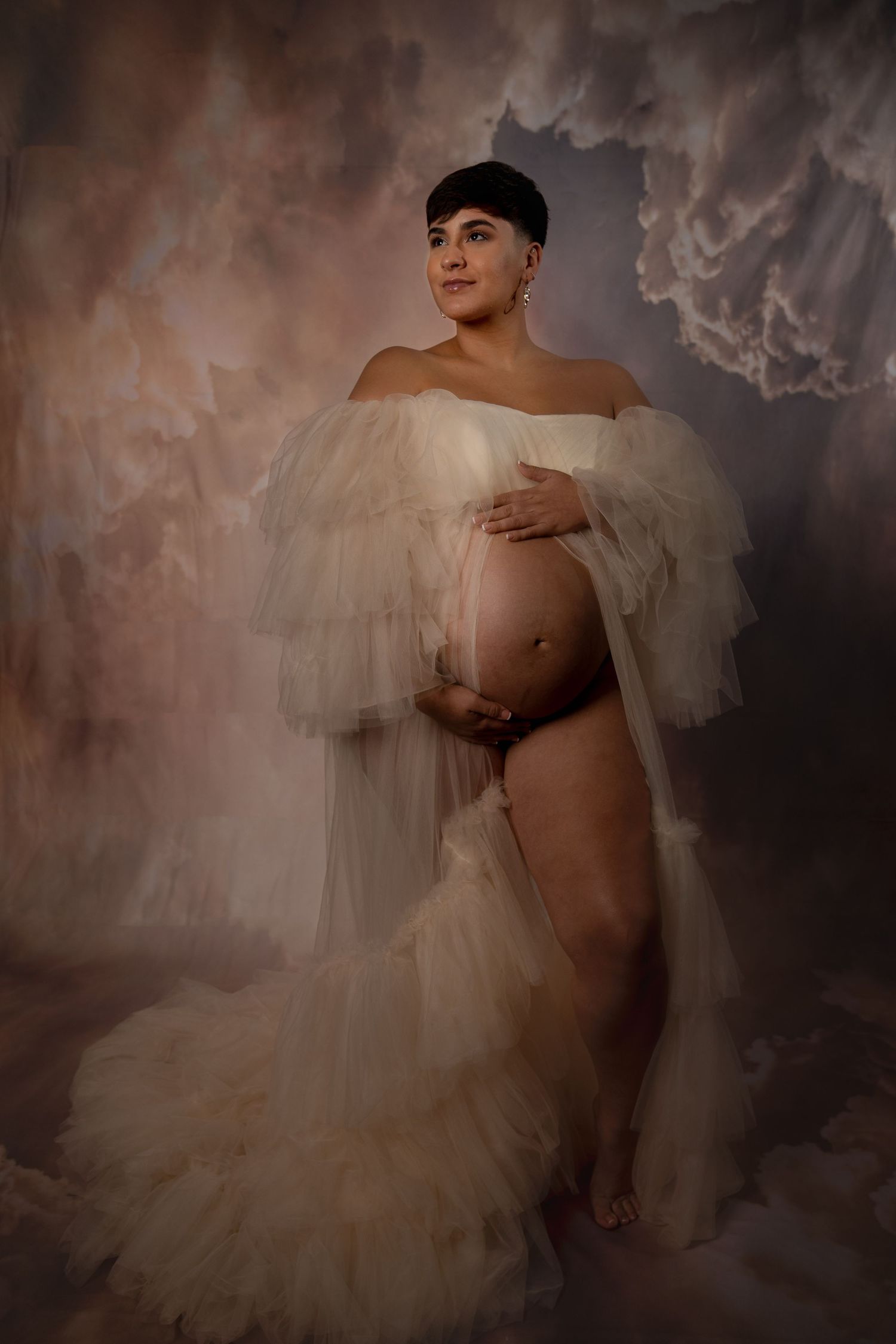 Ethereal maternity portrait with flowing white tulle dress against moody background.