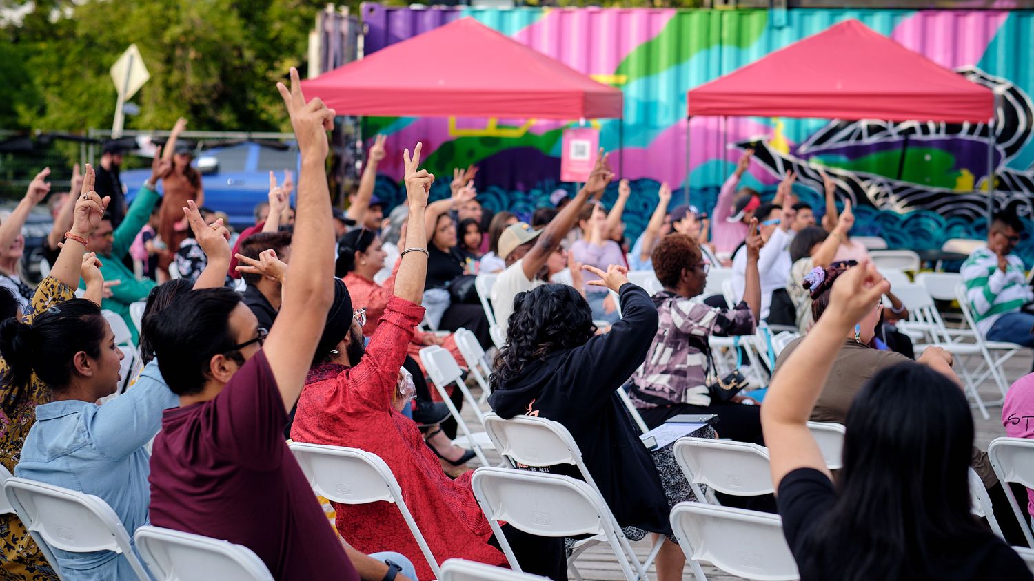 A crowd of people seated in white chairs raise their arms in celebration at an outdoor event with pink and blue decor.