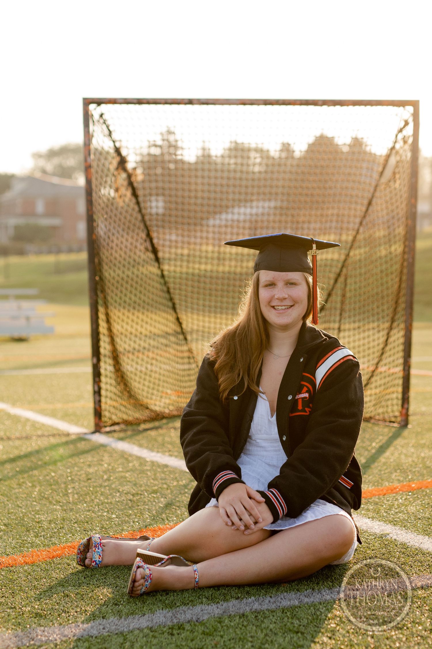 Graduate in varsity jacket sits on sports field with lacrosse goal in golden sunset.