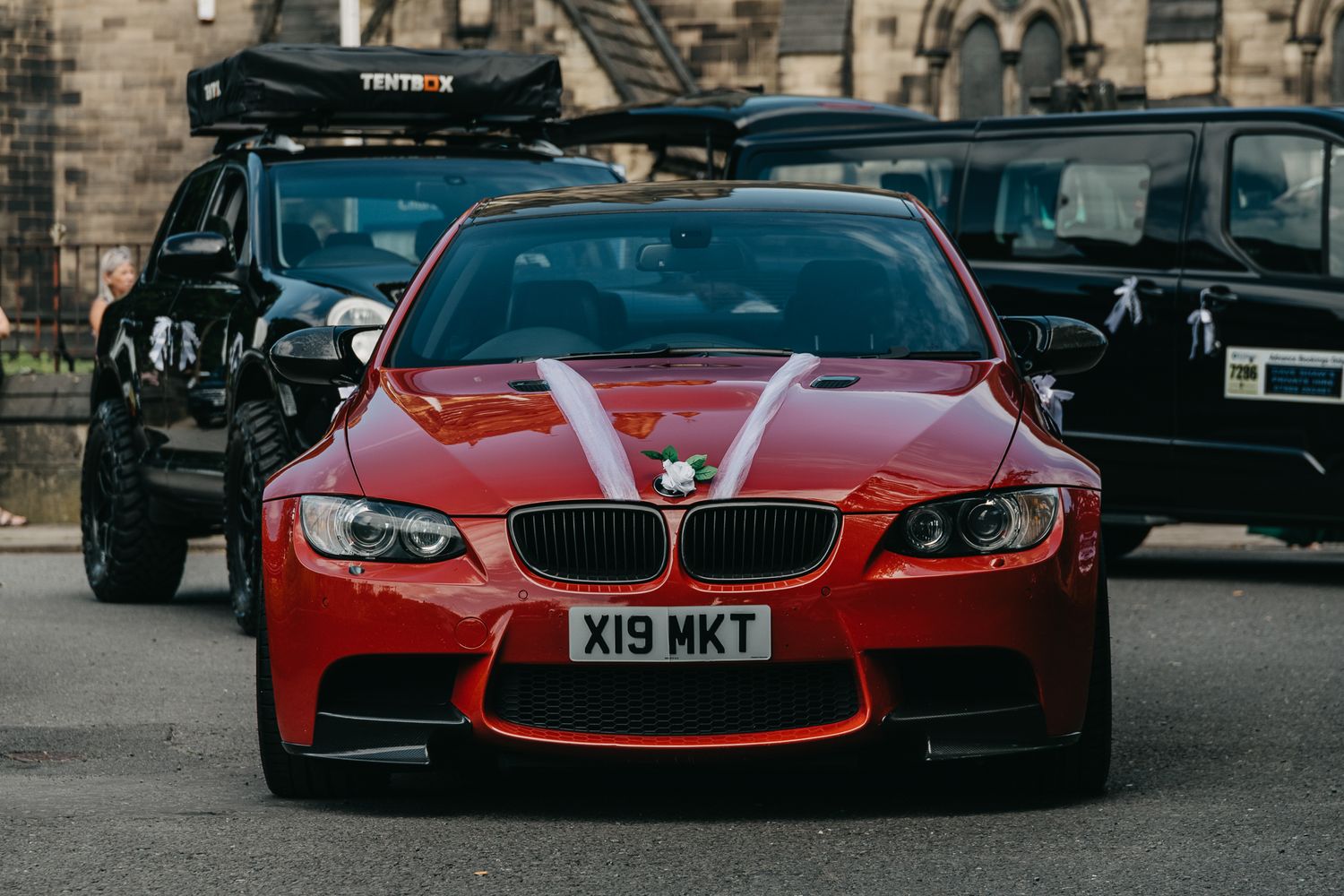 A red BMW M3 sports car with black wheels and a roof rack parked in front of a stone building.
