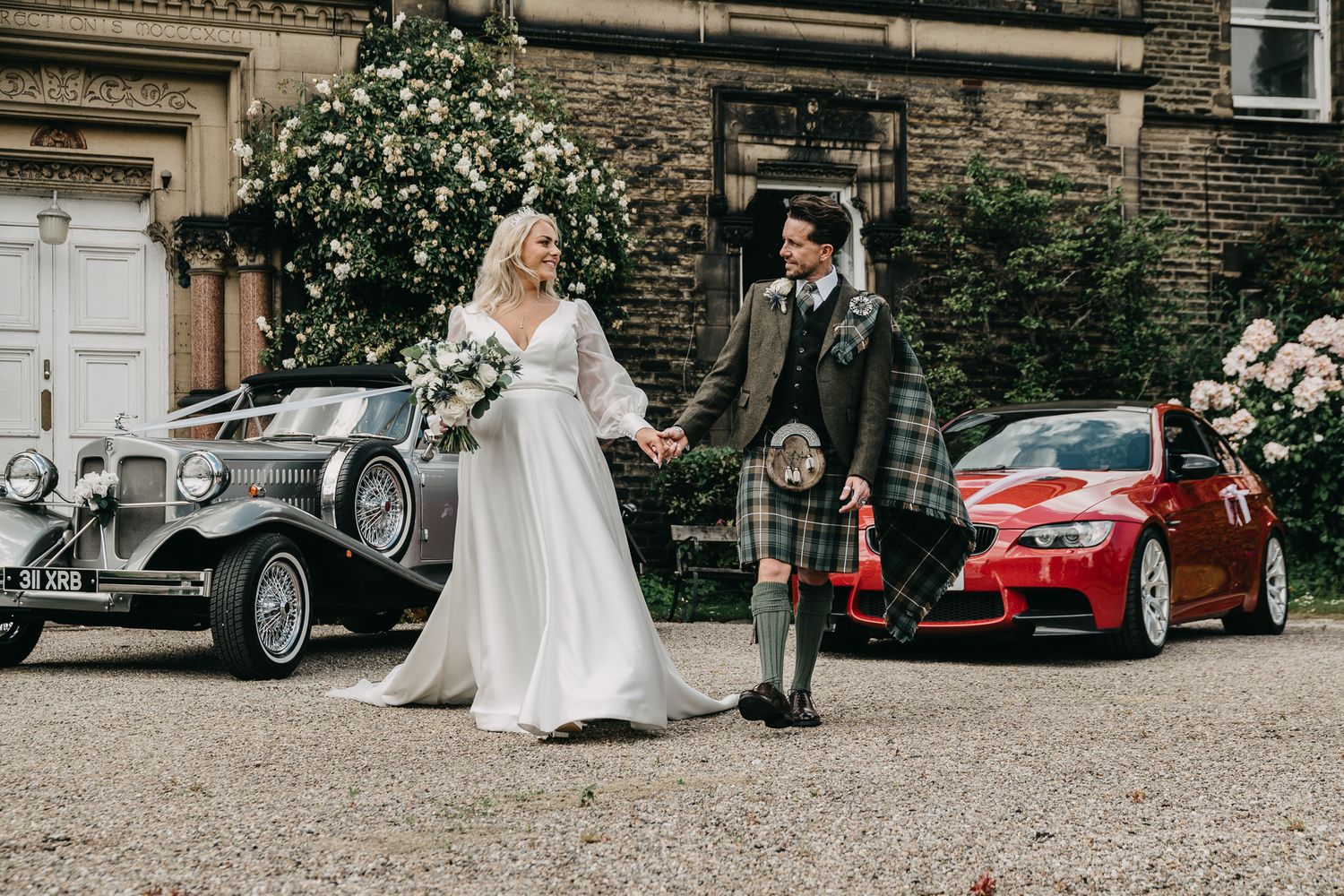 Classic vintage car parked alongside newlyweds in traditional Scottish attire outside stone building on wedding day.