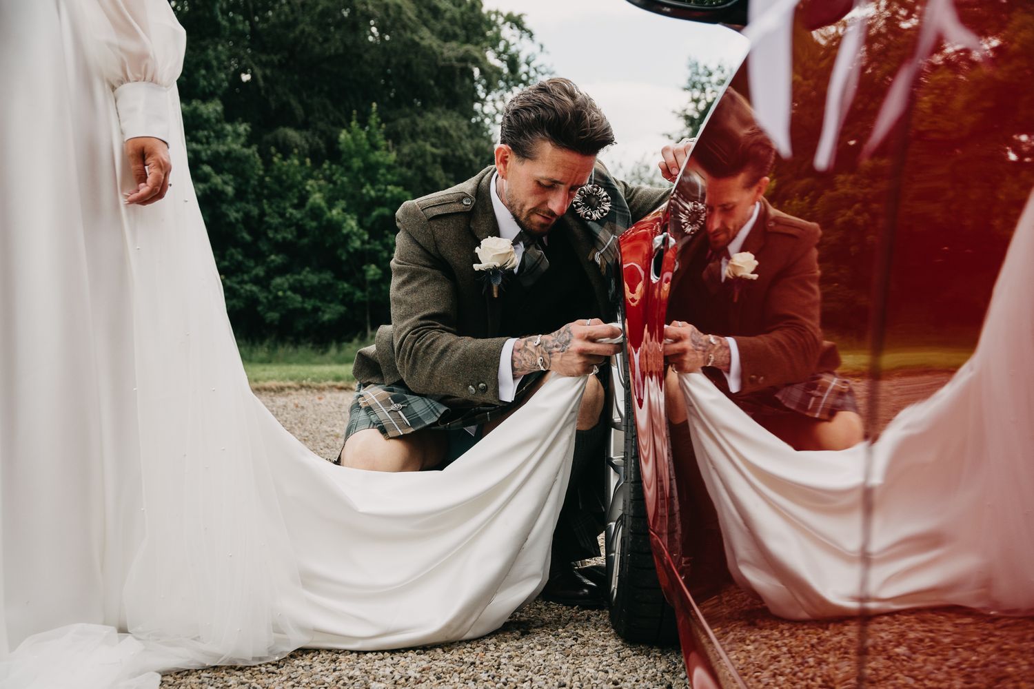 Someone in a brown jacket carefully arranges a white wedding dress near a red classic car outdoors.
