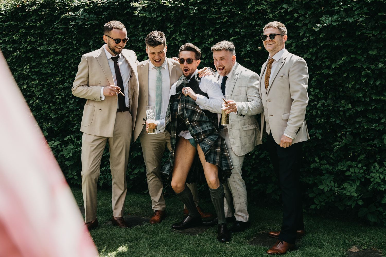 Wedding party in beige suits gather outdoors against green hedge backdrop.