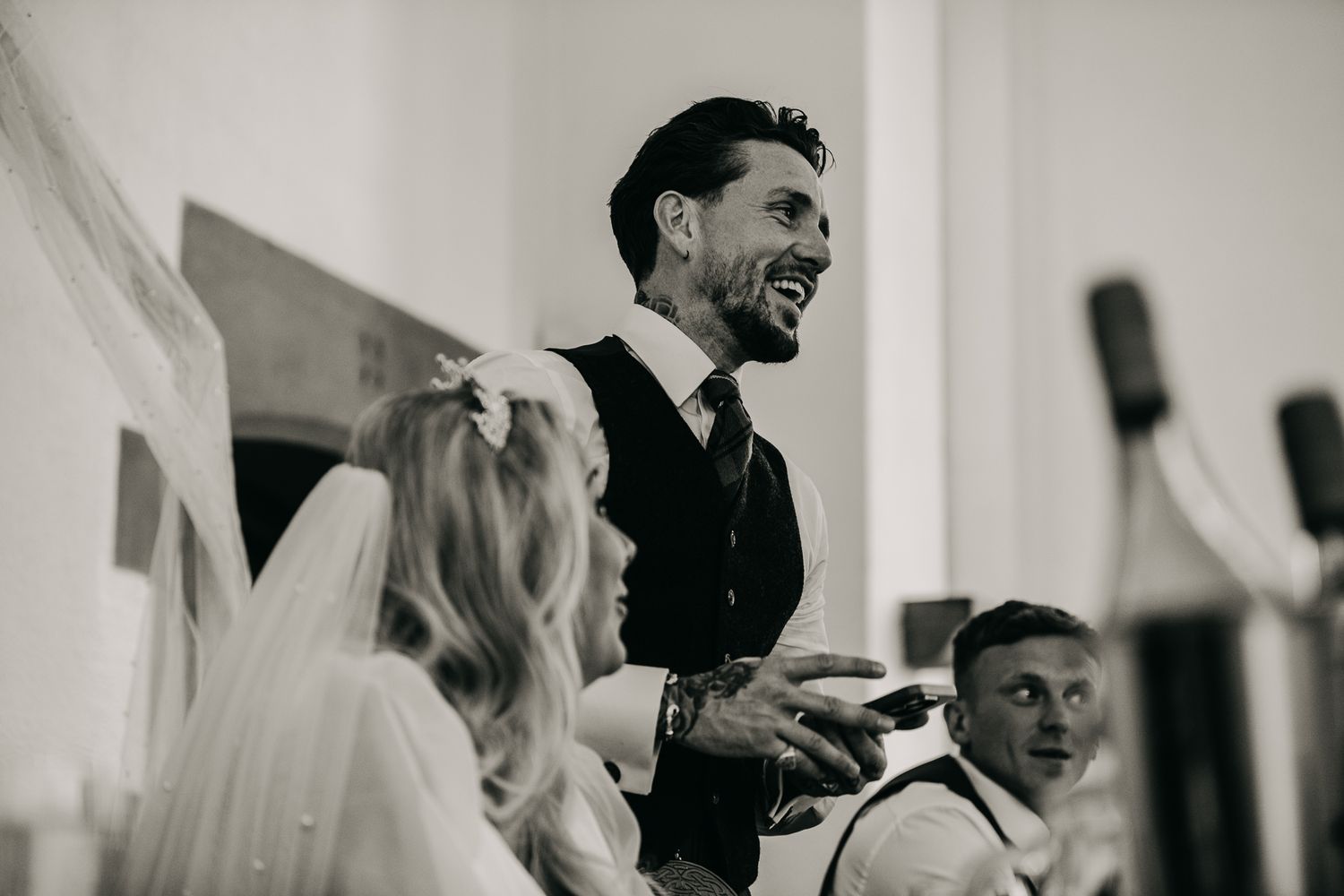 A candid black and white photograph captures joyful moments during wedding reception speeches.