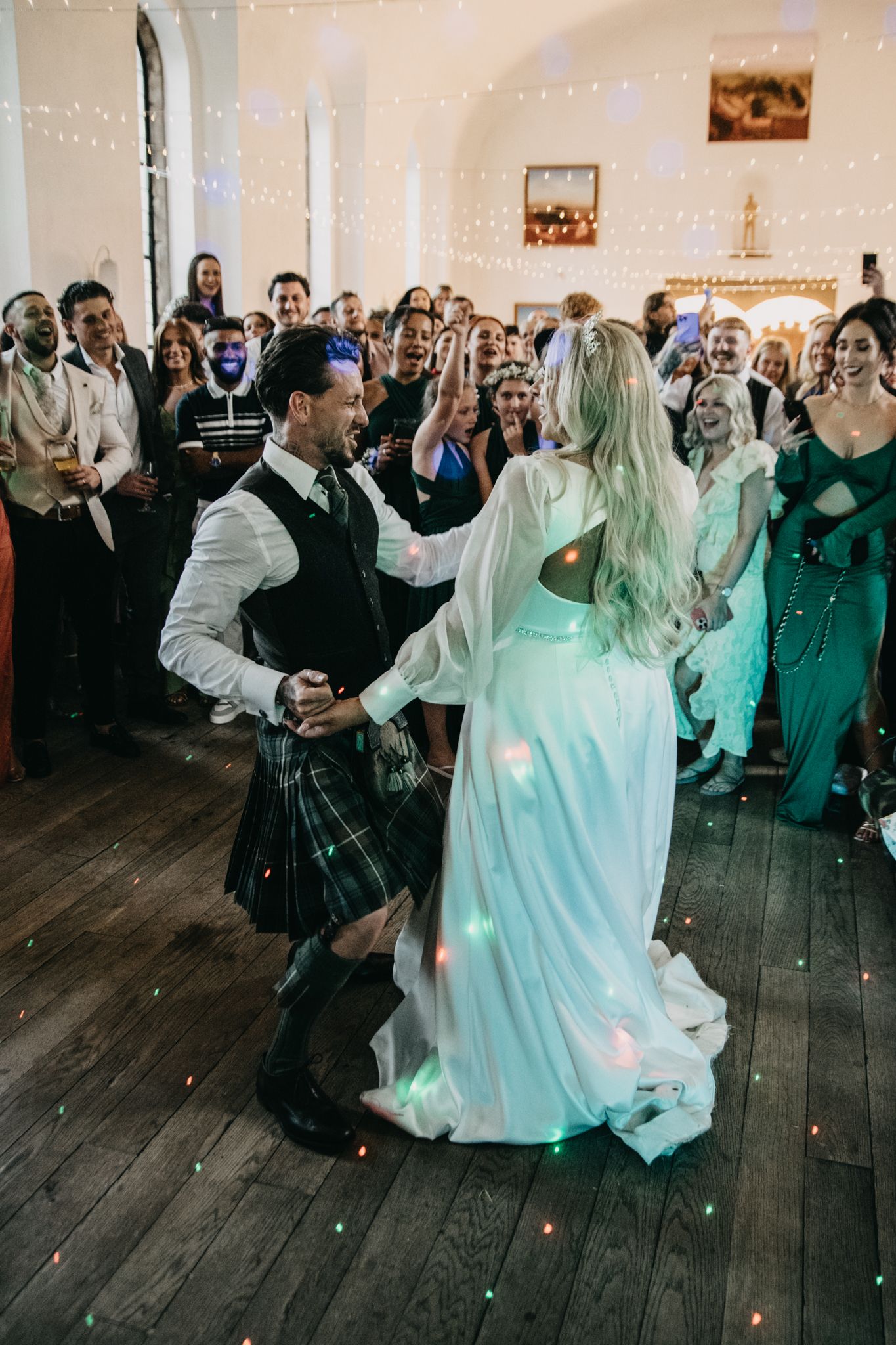 Couple performing first dance in traditional Scottish attire under twinkling lights.
