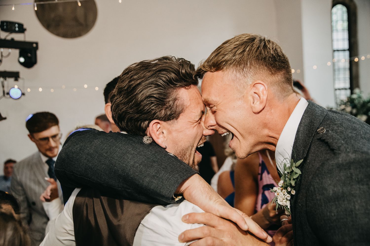 Two people share an emotional embrace and laugh during a wedding celebration indoors.