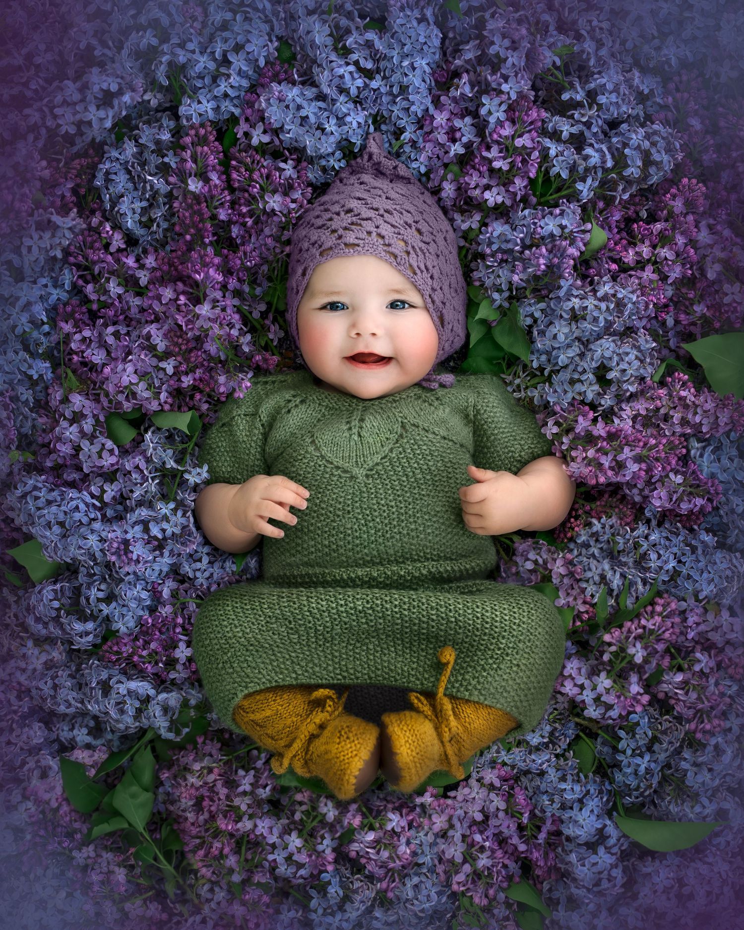 Smiling baby wearing green knit outfit surrounded by purple lilac blossoms.