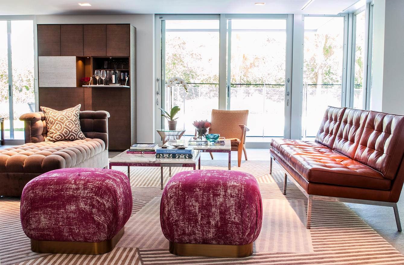 Modern living room with plush burgundy ottomans, brown leather bench, and large windows overlooking a scenic view.