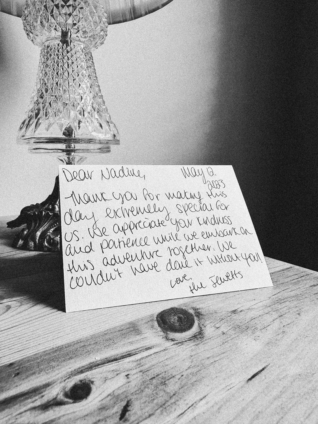 A handwritten thank you note displayed under a crystal light fixture on a wooden surface.