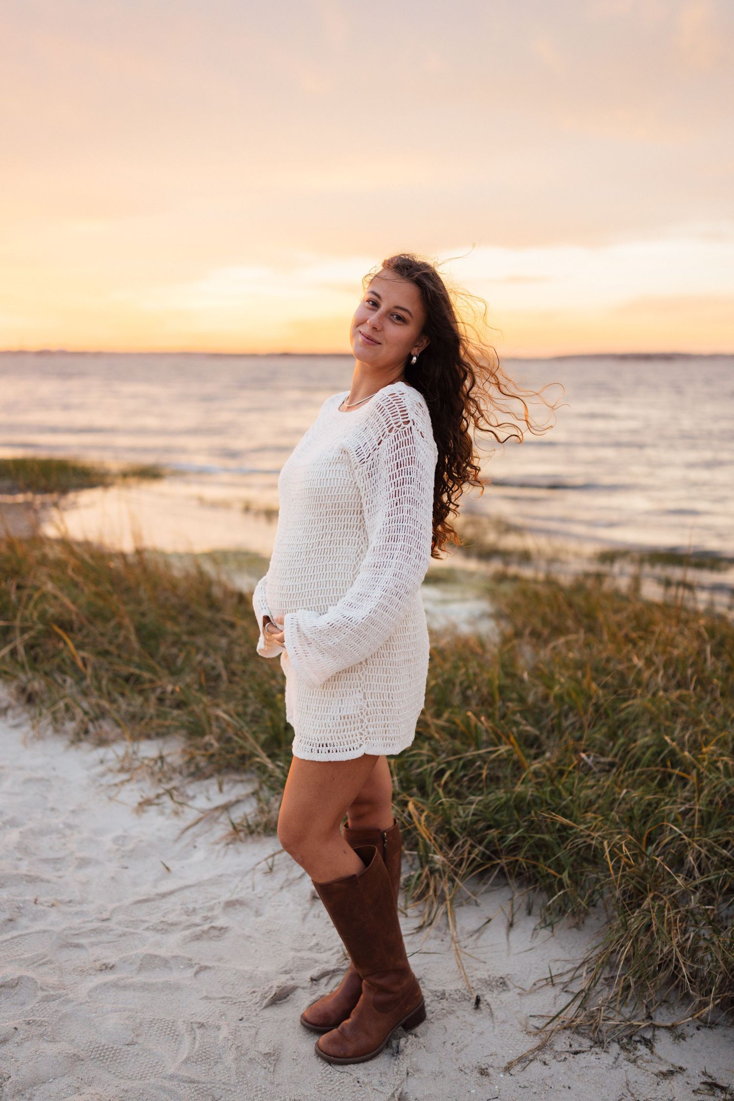 Woman in white crochet dress and brown boots poses on beach at sunset with windswept hair. Wilmington photographer.