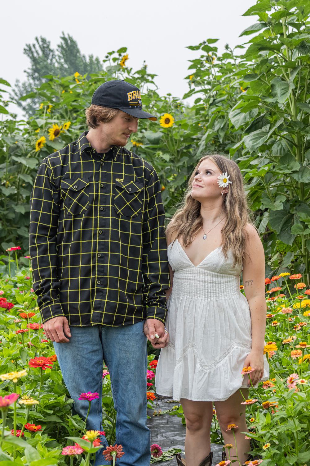 Couple holding hands in a colourful flower garden with sunflowers, woman in white dress with daisy in hair smiling at man. Man wearing a cap