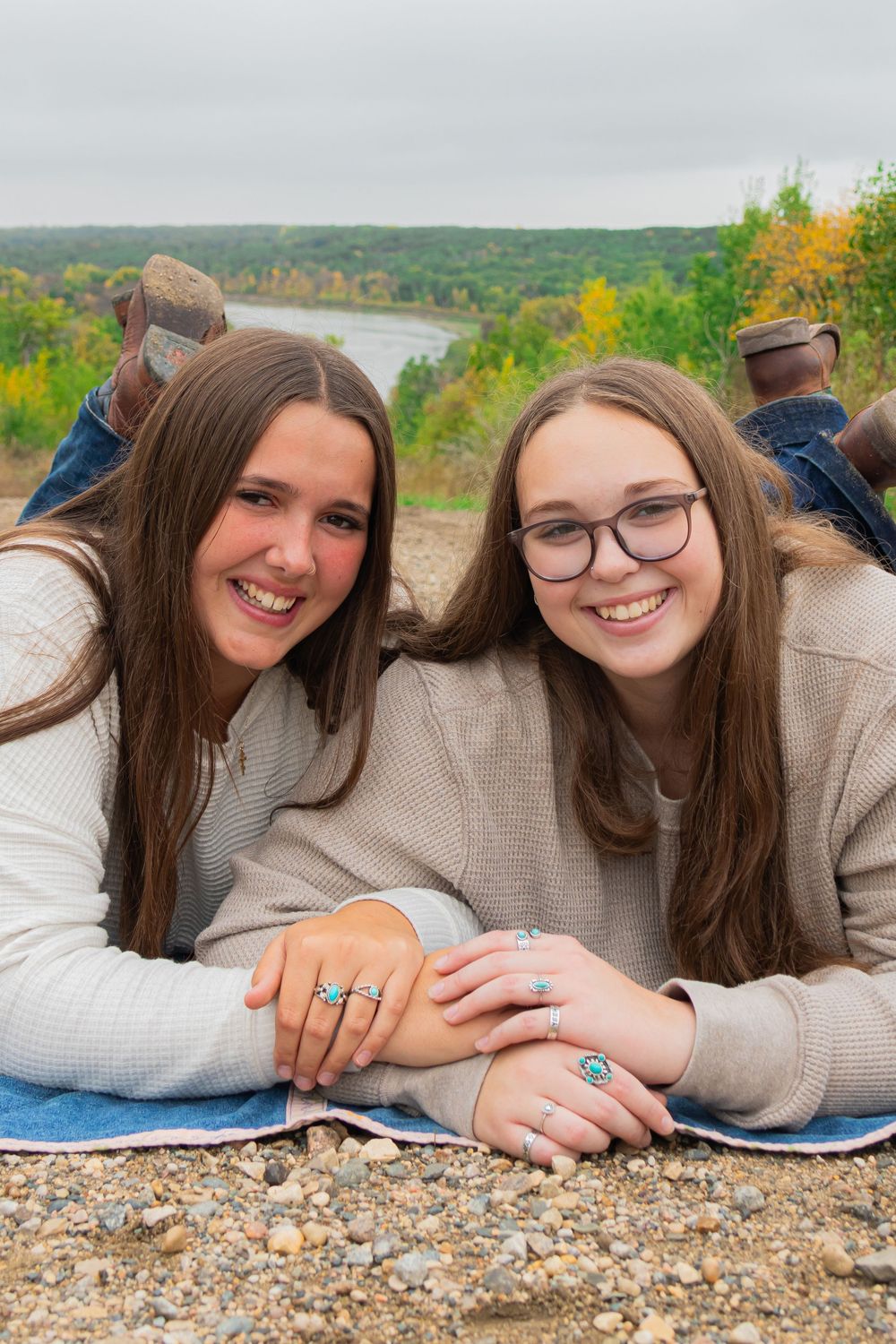 Two smiling teenage girls lying on a rocky overlook with autumn trees and a river in the background. Best friend photoshoot. Jewelry. Boots