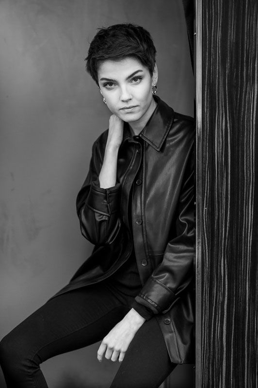 Woman with short pixie cut wearing oversized leather shirt poses thoughtfully in black and white studio portrait.
