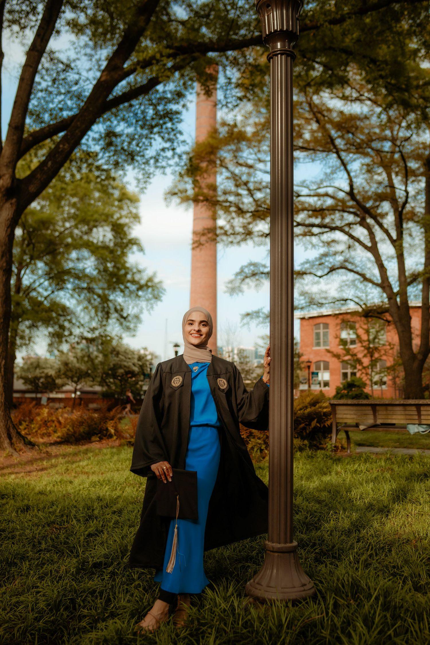 Graduate in black academic regalia poses near lamp post with red brick building in background.