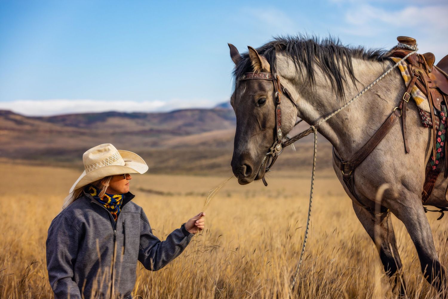 Cowgirl gestures while kneeling next to her young horse in vast grassland.