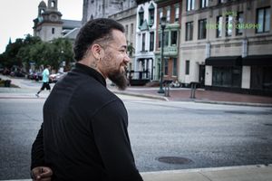 Person with beard in black clothing walking on urban street with buildings in background.