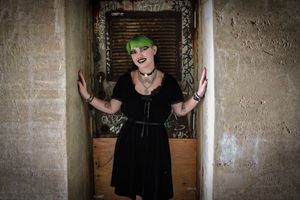 Alternative style person with green hair posing in gothic black dress against industrial doorway.