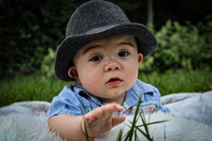 Baby wearing a gray fedora hat laying on a blanket outdoors.