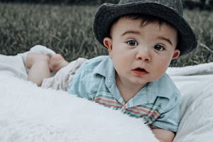 Baby in denim clothing and fedora hat resting on white blanket.