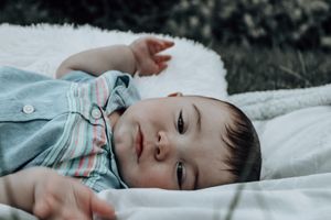 A small baby lying peacefully on a white blanket wearing a light blue outfit in a natural outdoor setting.