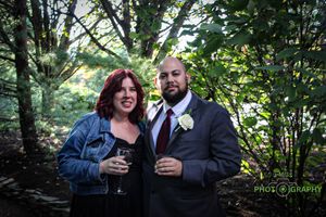 Couple in formal attire holding wine glasses in a wooded outdoor setting.