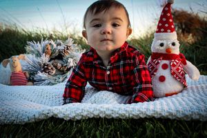 A winter holiday scene with a plaid-dressed baby and decorative snowman on a white blanket outdoors.