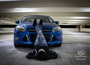 A bright blue Ford Focus parked in a concrete parking garage with someone laying on the ground in front of it.