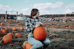 Person sitting with a large orange pumpkin at a pumpkin patch during autumn.
