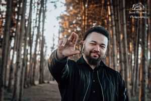 Person in a black jacket gesturing to camera in a forest during sunset.