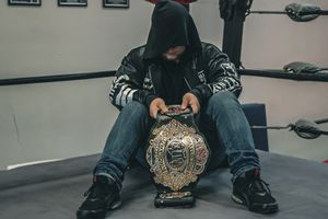 Person in black hoodie sitting with championship belt in a training facility.
