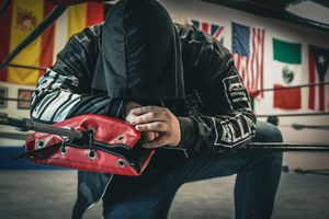 Person wearing a hooded jacket examining red boxing gear in a gym with international flags.