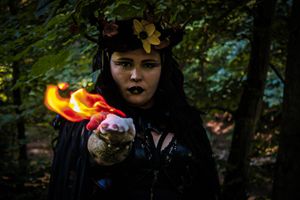 Dark figure in black attire surrounded by colored smoke effects in a wooded setting.
