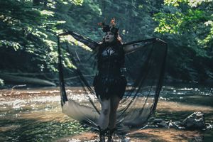 Silhouette with flowing black dress poses dramatically in sunlit forest clearing.