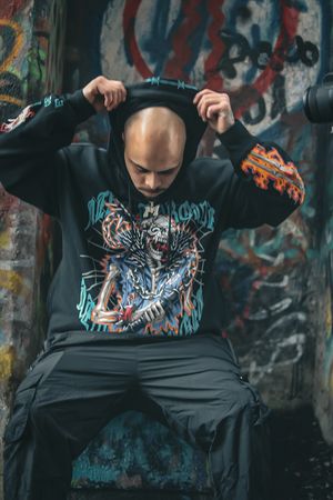 Person in graffiti-style hoodie poses against urban wall with street art.