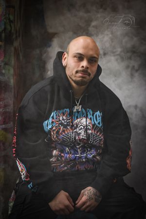 Person wearing graffiti-style hoodie poses in smoky urban setting with street art background.