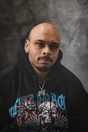 Close-up portrait of someone in a black hoodie with blue graffiti-style artwork against a smoky background.