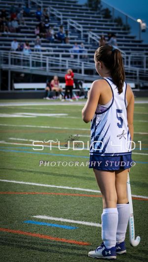 Field hockey player kneels on artificial turf field wearing white uniform during practice or game.