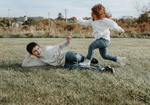 Two playful people in casual clothing laughing while playing and falling on grass in a field on a sunny day.
