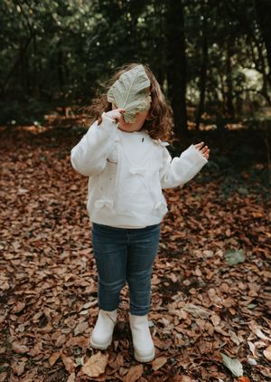A small child in a white sweater and jeans explores autumn leaves scattered on the forest floor.