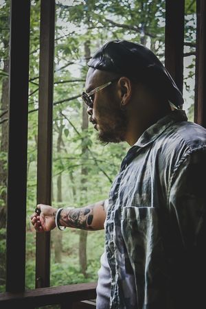 Person with tattoos and sunglasses looking to the side while standing on a forest deck.