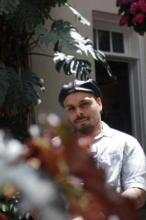 Person wearing baseball cap sits outdoors with foliage and white building in background.