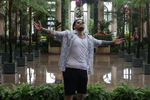Person in a greenhouse wearing a white shirt and black pants surrounded by lush tropical plants and flowers.